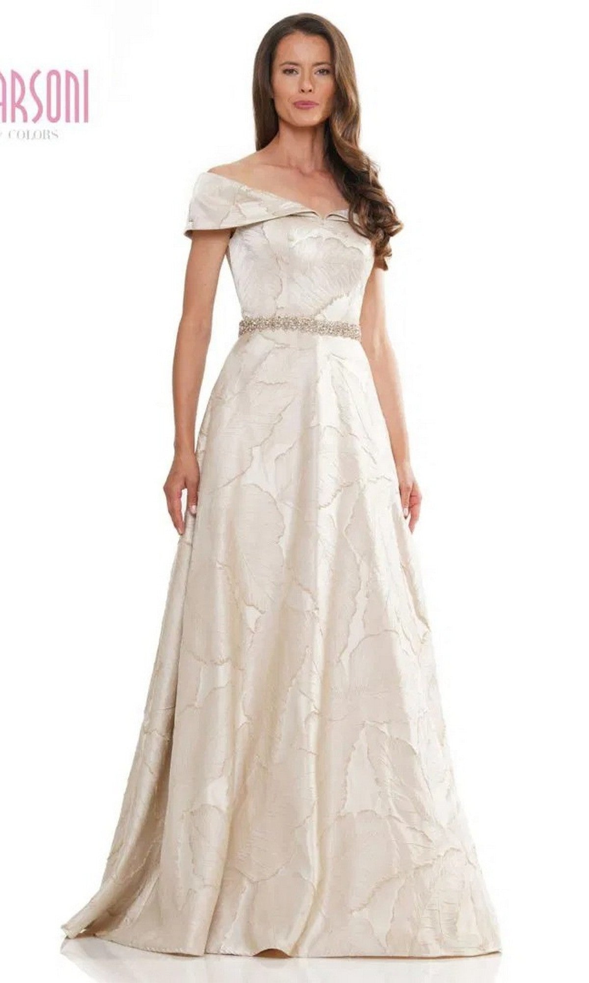 A woman stands against a white background wearing the Marsoni MV1265 long formal dress in champagne, featuring an off-the-shoulder neckline, textured pattern, and jeweled belt for an elegant touch.